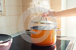 Female hand open lid of enamel steel cooking pan on electric hob with boiling water or soup and scenic vapor steam