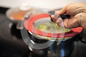 Female hand open pan lid on electric hob with boiling water or green soup and scenic vapor steam backlit.