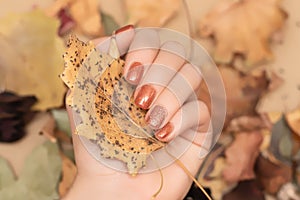Female hand with orange autumn nail design. Woman hand hold dry leaf. Autumn nail polish manicure with golden glitter
