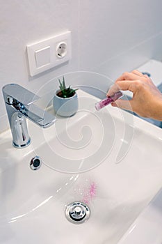 Female hand pouring purple glitter bottle into washbasin at bathroom and polluting clean water