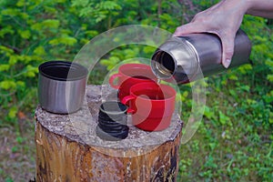 A female hand pours tea from a thermos into cups standing on a stump