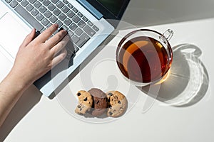 Female hand prints on a laptop, a cup with tea, and three chocolate chip cookies on a white background in sunlight