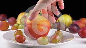 Female Hand Putting a Ripe Apple or Peach on a Plate with Grape and Fruit