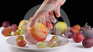 Female Hand Putting a Ripe Apple or Peach on a Plate with Grape and Fruit