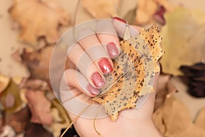 Female hand with red autumn nail design. Woman hand hold dry leaf. Autumn nail polish manicure with golden glitter