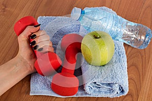 Female hand with red dumbbell and fresh green apple and water bottle on blue towel on wooden floor