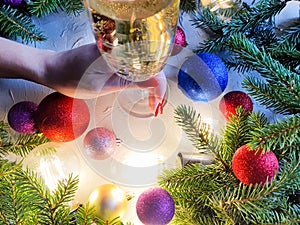 Female hand with red nails with glass of champagne, branches of christmas fir tree, magic lights and shiny balls on gray table