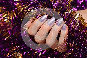 Female hand with silver Christmas nail design. Silver nail polish manicure. Female hand holding purple New Year tinsel