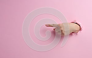 Female hand sticks out of a torn hole in a pink paper background, part of the body points with the index finger to the side