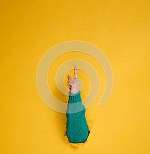 Female hand sticks out of a torn hole in a yellow paper background, part of the body points with the index finger to top. Copy