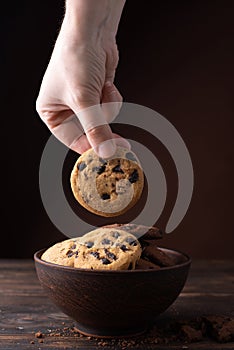 A female hand takes one chocolate chip cookie from a plate on a dark woody background