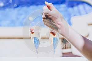 Female hand throws ice into a glass champagne with strawberry on sunny terrace overlooking swimming pool at summer day