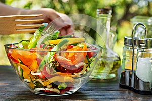 Female hand tucks a full bowl of fresh vegetable salad on a wooden table outdoors against a background of blurred green leaves