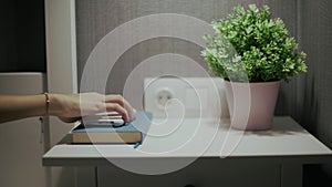 Female hand turn off an alarm clock on mobile phone on bedside table with indoor plant and book