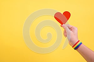 Female hand wearing rainbow wristband holding red heart shaped paper  on yellow background. LGBT, Pride, LGBTQ Symbols