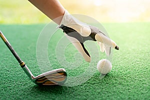 Female hand wearing white professional gloves with the left hand putting golfball and the putter golf on the grass course