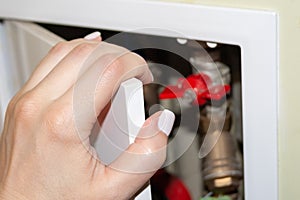 A female hand with white nails opens a white door in the recess of the bathroom wall, behind which pipes, counters and valves