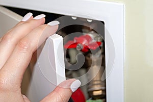 A female hand with white nails opens a white door in the recess of the bathroom wall, behind which pipes, counters and valves