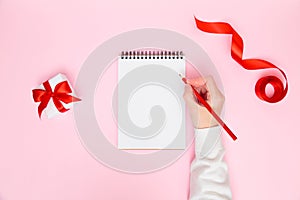 Female hand writing in blank notebook with festive decorations, gift box on pastel pink background. New year planning