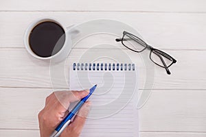 Female hand are writing in a blank notebook on a white wooden background. Glasses and a cup of coffee. Flat lay