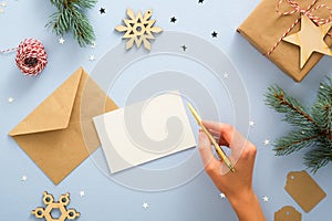 Female hand writing letter to Santa Claus over blue background with festive Christmas decorations, confetti, fir tree branches,