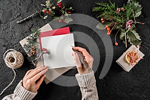 Female hand writing a letter to Santa on dark background with Christmas gift, berries, Fir branches, skein of jute. Xmas