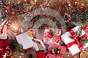 Female hand writing a letter to Santa on holiday background with Christmas gifts, candle, Fir branches.