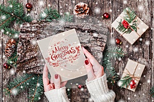 Female hand writing a letter to Santa on wooden background with Christmas gifts, bark texture, Fir branches, pine cones, red decor