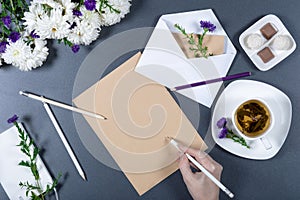 Female hand writing on a sheet of brown craft paper. Fresh chrysanthemums, pencils, envelope with note and flower, cup of tea and