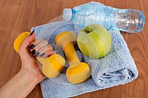 Female hand with yellow dumbbell and fresh green apple and water bottle on blue towel on wooden floor