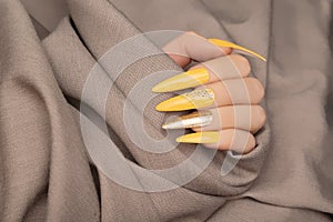 Female hand with yellow nail design. Long nail polish manicure. Woman hand on beige fabric background