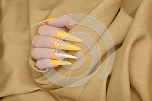 Female hand with yellow nail design. Long nail polish manicure. Woman hand on beige fabric background