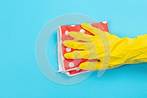 A female hand in a yellow rubber protective glove with a micro fiber red rag wiping a blue wall from dust. A maid or housewife