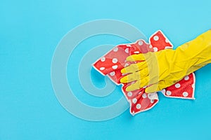 A female hand in a yellow rubber protective glove with a micro fiber red rag wiping a blue wall from dust. A maid or housewife