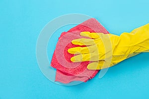 A female hand in a yellow rubber protective glove with a micro fiber red rag wiping a blue wall from dust. A maid or housewife