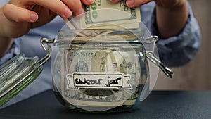 Female hands add money to Glass jar full of American currency dollars cash banknote with text SWEAR JAR. Preparation