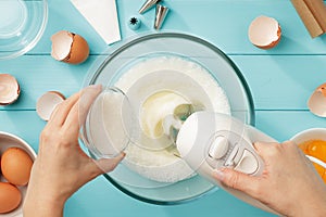 Female hands add a sugar to the bowl with whipping egg whites with mixer.