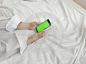 Female hands on a background of a bed holding a phone with a green screen