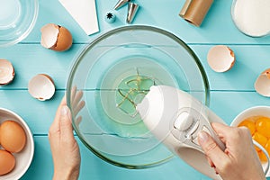 Female hands beating egg whites cream with mixer in the bowl on blue wooden table.