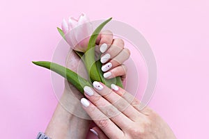 Female hands with beautiful manicure - white ivory oval nails with tulip flower on pink paper background, closeup