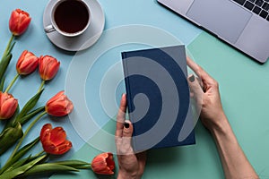 Female hands with book on blue background. Background with coffee, laptop and flowers.