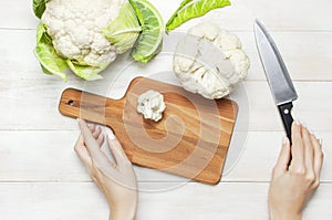 Female hands cut cauliflower with knife on cutting board, kitchen towel on rustic white wooden background top view flat lay copy