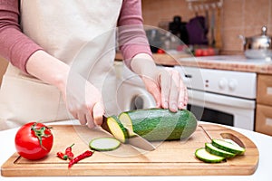 Female hands cut with a knife a young seasonal zucchini into slices on a wooden cutting board. The method of preparation of