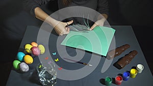 Female hands cut into thin fringe the green paper with scissors over gray table.