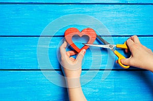 Female hands cut a wooden red heart with scissors on a blue background. The concept of breaking relations, quarrel, divorce. The p