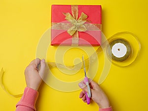Female hands are cutting a silk ribbon with scissors, next to gift boxes. Yellow background, top view