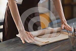 Female hands flatting piece of clay lying on fabric using wooden rolling pin in ceramic studio.