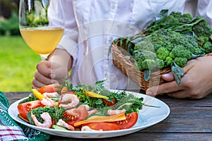 Female hands with a glass of wine hold a basket with dill, next to it there is a vegetable salad with shrimps in a plate on the