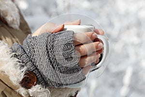 Female hands in gray knitted mittens with cup of tea at background of snowy forest