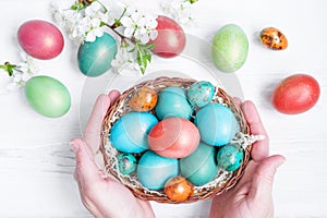 Female hands hold a basket of bright Easter eggs decorated with spring flowers on a white wooden surface  top view.